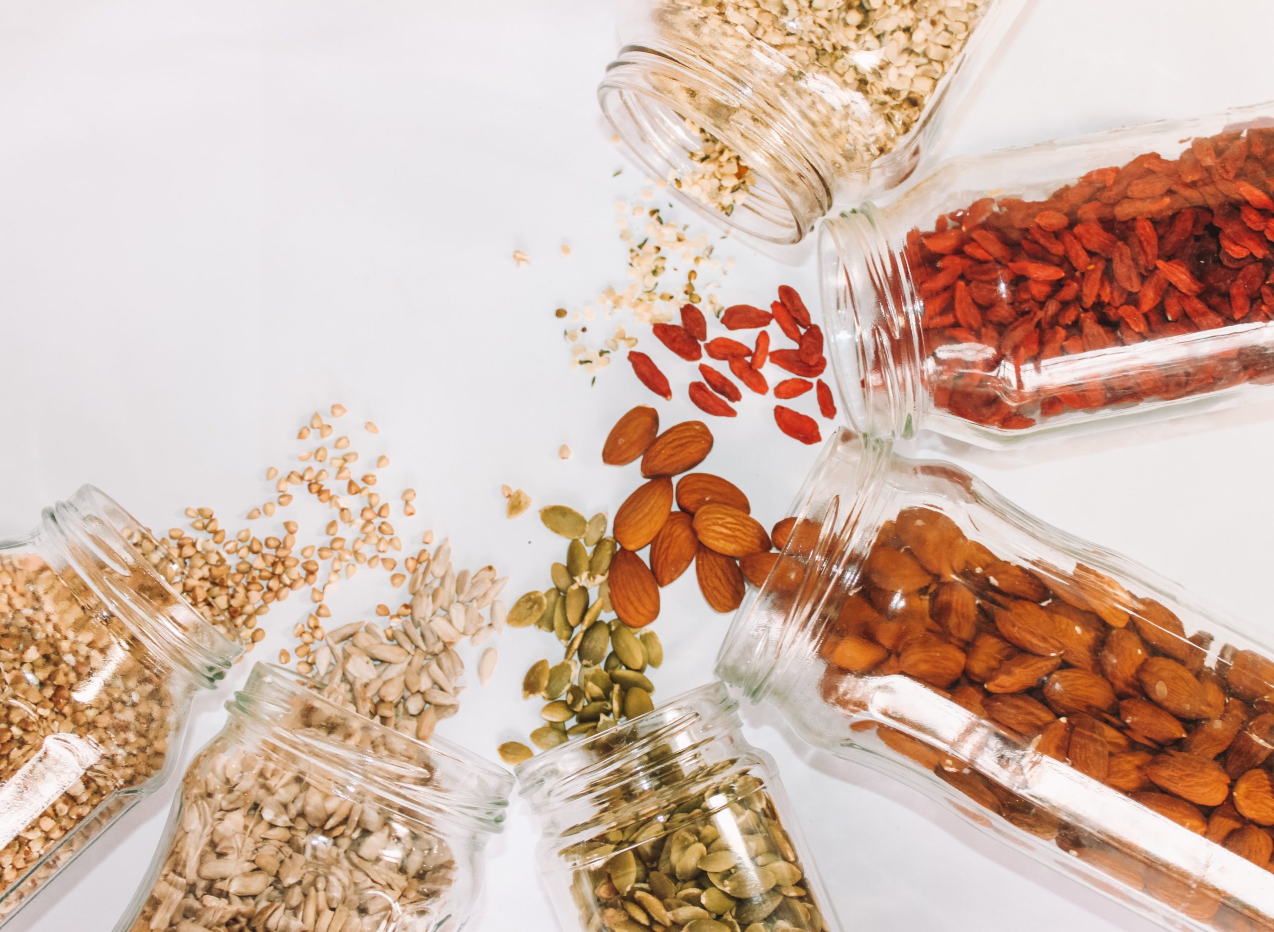 five glass jars in a semi-circle. each pouring out a different nut, seed, or grain. almonds, pumpkin, sunflower seeds, goji berry, barley, and one other tan seed or nut. white background