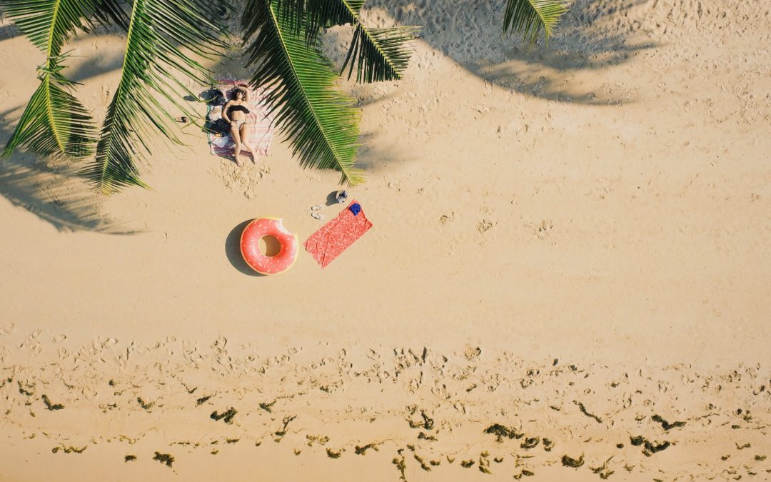 woman lying on a beach, aerial view. Green palm fronds and pink floatie with a pink towel lie on the sand